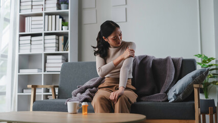 Fototapeta premium Young woman is experiencing a painful shoulder while sitting on her couch at home with a bottle of pills and a mug on the table in front of her