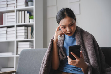 Naklejka premium Sick young woman with a fever wrapped in a blanket, holding a tissue and checking symptoms on a smartphone while sitting on a sofa at home