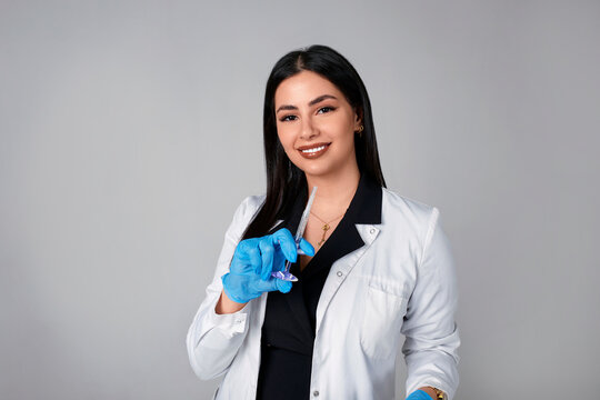 Portrait of an American beautiful female cosmetologist doctor in a white coat with an injection syringe. Medical student. Female hospital worker looking at camera and smiling, studio, gray background - Powered by Adobe