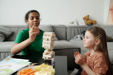 Side view of cute little child sitting in front of table and looking at wooden block tower built by African American teenage baby sitter