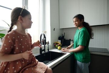 Happy teenage girl in t-shirt and jeans looking at adorable child sitting on kitchen counter while chopping fresh vegetables on plastic board