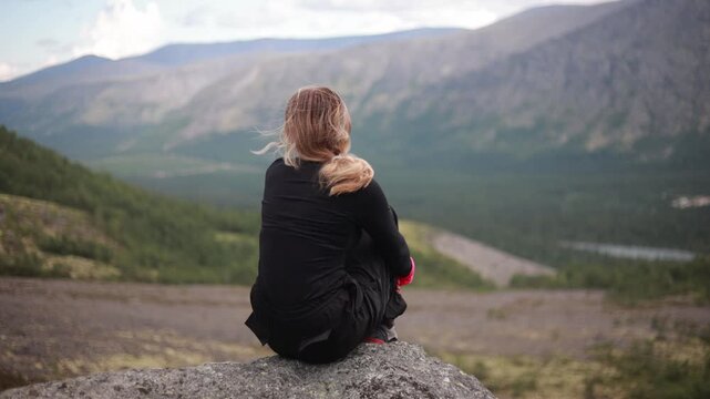 A young woman sits on a rock and admires the mountain valley
