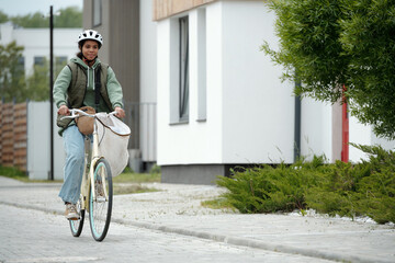 Fototapeta premium Cute African American teenager in casualwear and protective helmet sitting on bicycle with white sack containing newspapers and other post
