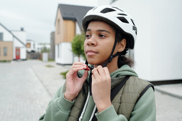 Cute African American teenage girl putting on protective helmet and fastening it under neck while preparing for delivery of post