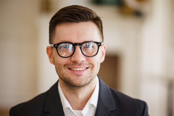 A smiling businessman in glasses and a suit looks directly at the camera in a bright office.