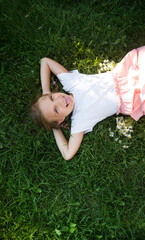 A portrait laughing  child girl l in a dress with pockets in which daisy bouquet  in her pocket when looking at camera lies on the green grass on a summer day.
