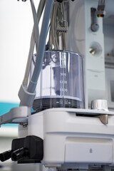 Medical Equipment With Graduated Container. A close-up of medical equipment with a graduated container, showing a portion of the machine's white casing, and a white knob.