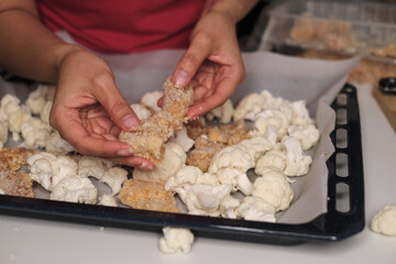 Hands arranging fresh cauliflower florets and breaded chicken on a parchment-lined baking tray, capturing the detailed process of meal preparation. Reflects the modern trend of healthy, home-cooked