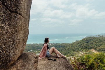 A woman sits on a rocky cliff overlooking the ocean and lush green jungle in Thailand