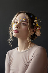 Naklejka premium A young woman with blonde hair, wearing a plain long-sleeve shirt, gazes upwards as she poses in front of a dark background. She has flowers in her hair and on her face