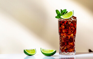 Rum cola cocktail long drink with ice, mint and lime in glass. Beige background