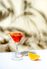 Orange cocktail with red vermouth, liqueur, bitter, citrus zest and ice in martini glass on beige background, hard light