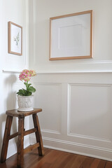 A close-up photo of white wainscoting in an interior, with a picture frame hanging above it. The floor is wooden and there's some greenery on top of one stool.