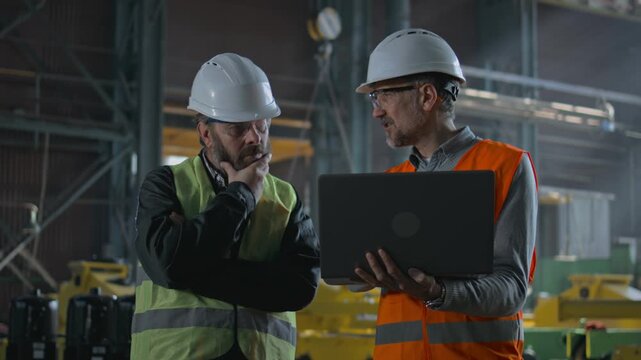 Two professional heavy industry workers, engineers wearing protective uniform and hardhats discuss work process using laptop computer while standing in manufacturing factory or warehouse. Slow motion.