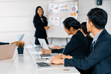 Asian business people listening to presentation from businesswoman who stands in front of group meeting room at international business conference. Multicultural corporate strategy concept. uds