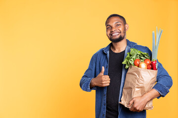 Smiling pleased male model gives shows thumbs up symbol in studio, posing with his groceries paper...