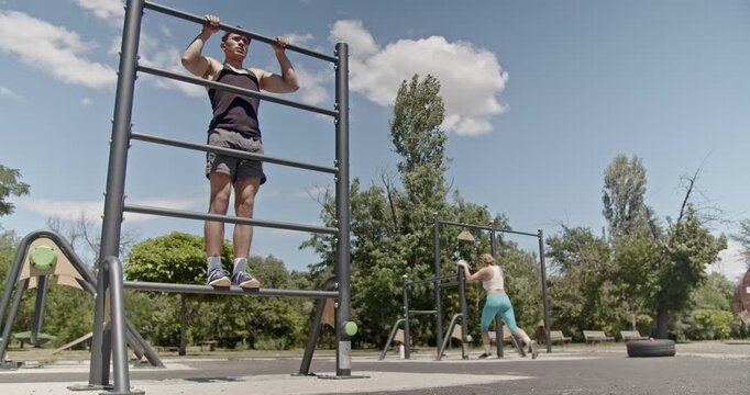 Fit sports couple exercising on outdoor gym equipment in a park on a sunny day. Sports man exercising his calves while standing on parallel bars.