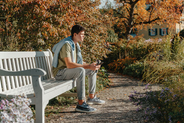 A Teenager Sits On A Bench In The Autumn Park Drinks Coffee From A Thermo Mug And Looks Into A Phone. Portrait Of Handsome Cheerful Guy Sitting On Bench Fresh Air Drinking Latte 
