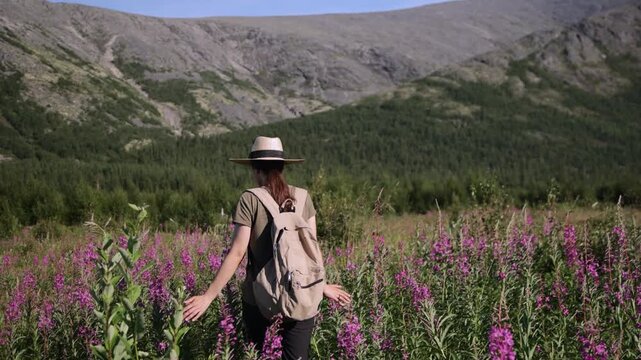 A girl in a straw hat walks through a mountain valley among flowers