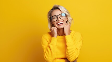 Happy blonde woman with round glasses and casual sweater, smiling with hands together. Vivid yellow background enhances her joyful expression, conveying positivity and friendly demeanor