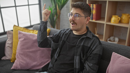 A young hispanic man with a moustache, wearing glasses, sits on a sofa indoors, gesturing upwards with an idea expression.