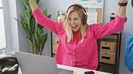 Blonde woman celebrating with arms raised in a modern office wearing headphones