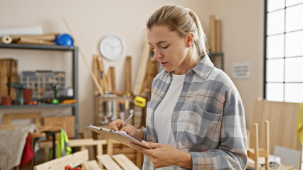 Focused woman writing on clipboard in a sunlit carpentry workshop