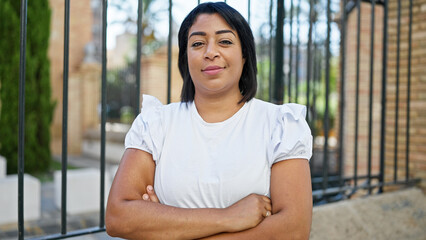 Confident hispanic woman with crossed arms in urban park setting, clear sky, greenery around.