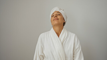 Beautiful mature hispanic woman in a white robe and head towel smiling with eyes closed against a white background