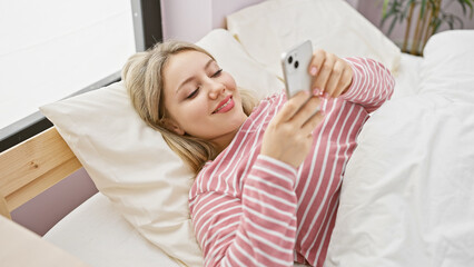 Obraz premium A young blonde woman in striped pajamas enjoys leisure time with her smartphone in a cozy bedroom setting at home.