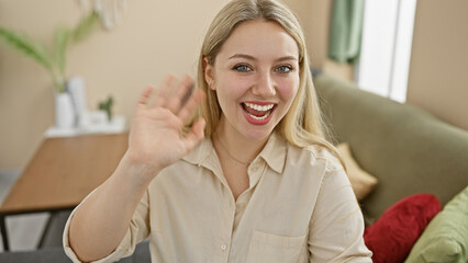 A cheerful young caucasian woman is waving hello in a bright cozy indoor home setting.