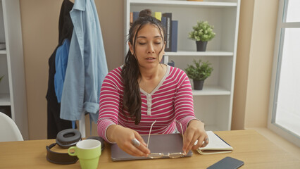 A young hispanic woman wearing a striped shirt cleans her glasses at a home office desk, depicting a casual indoor setting.