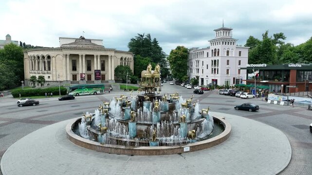 Aerial view of square with roundabout intersection with Colchis fountain in the middle of it filmed by drone in Kutaisi city. Cars are driving around it along buildings.