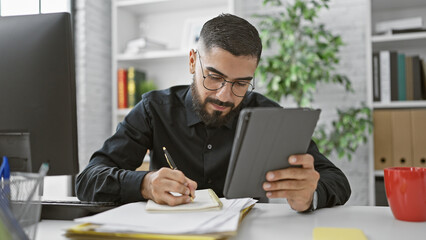 A focused man with a beard writing in a notebook and checking a tablet in a modern office setting