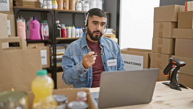 A bearded man volunteers at a donation center, managing inventory on a laptop in a warehouse setting.