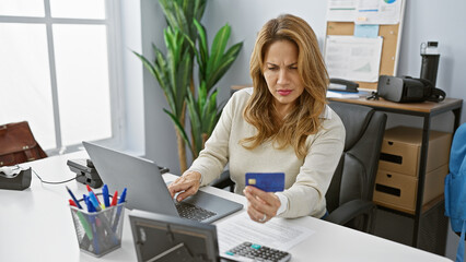 Focused hispanic woman analyzing finances with credit card in modern office setting