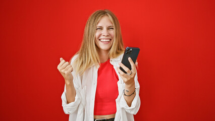 Blonde woman celebrating with a smartphone against a vibrant red background, expressing happiness and success.