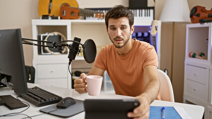 Handsome young man holding coffee mug at podcast setup in home music studio with microphone and instruments