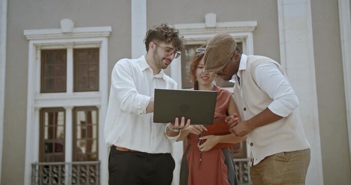 A diverse team of professionals engages in a project discussion outside, using a laptop. Their interaction highlights the elements of teamwork, collaboration, and modern business technology.