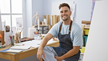 Handsome hispanic man with beard in denim apron sitting at a drawing table in an art studio