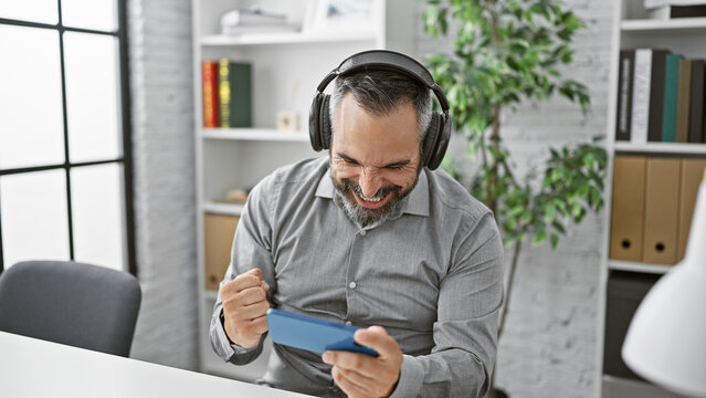 A cheerful bearded senior man with headphones plays a video game in a modern office, showcasing excitement and leisure during a break.