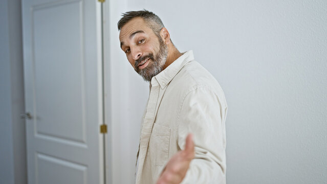 A charming mature man with grey beard welcomingly gestures at the house entrance.