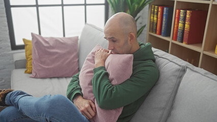 Fototapeta premium A thoughtful bald man with a beard embracing a cushion on a sofa in a cozy living room setting.