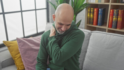 Fototapeta premium Bald handsome hispanic man with beard feeling back pain at home, sitting on a gray couch in a living room.