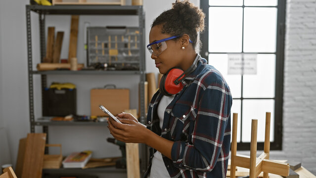 African american woman in safety glasses using smartphone in carpentry workshop