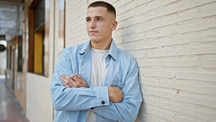 Handsome young hispanic man with arms crossed leaning against a brick wall on the urban city street.