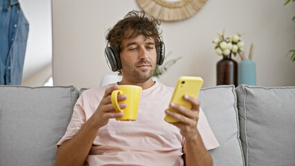 Handsome hispanic man relaxing with coffee and smartphone on couch in cozy home interior