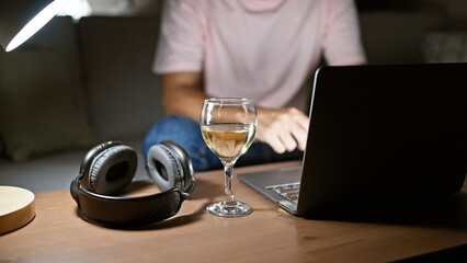 A relaxed man works from home, his laptop, wine glass, and headphones on a wooden table against a cozy indoor background.