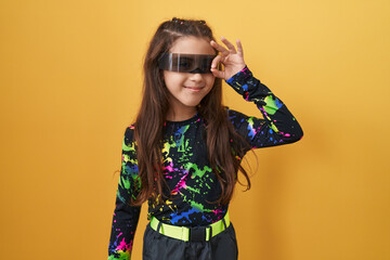 Adorable hispanic girl bursting with joy, sporting virtual reality glasses, cheekily doing the ok sign with finger through eye. all smiles against a sunny yellow isolated background.