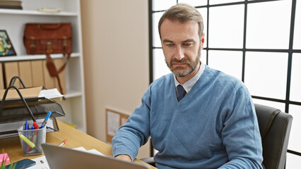 Focused middle-aged hispanic man with beard working on laptop in bright office interior.